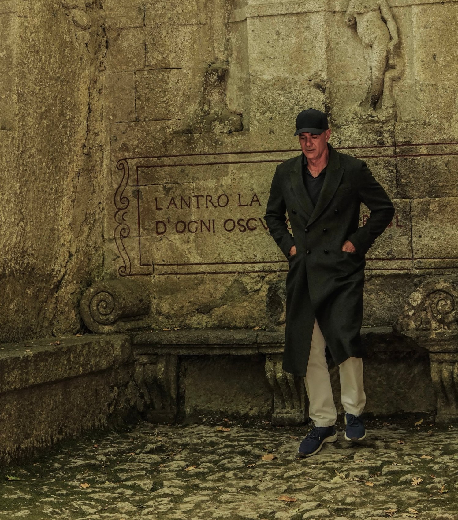 Andrea Provvidenza in a long dark double-breasted loden coat and a black cap, walking past a stone wall