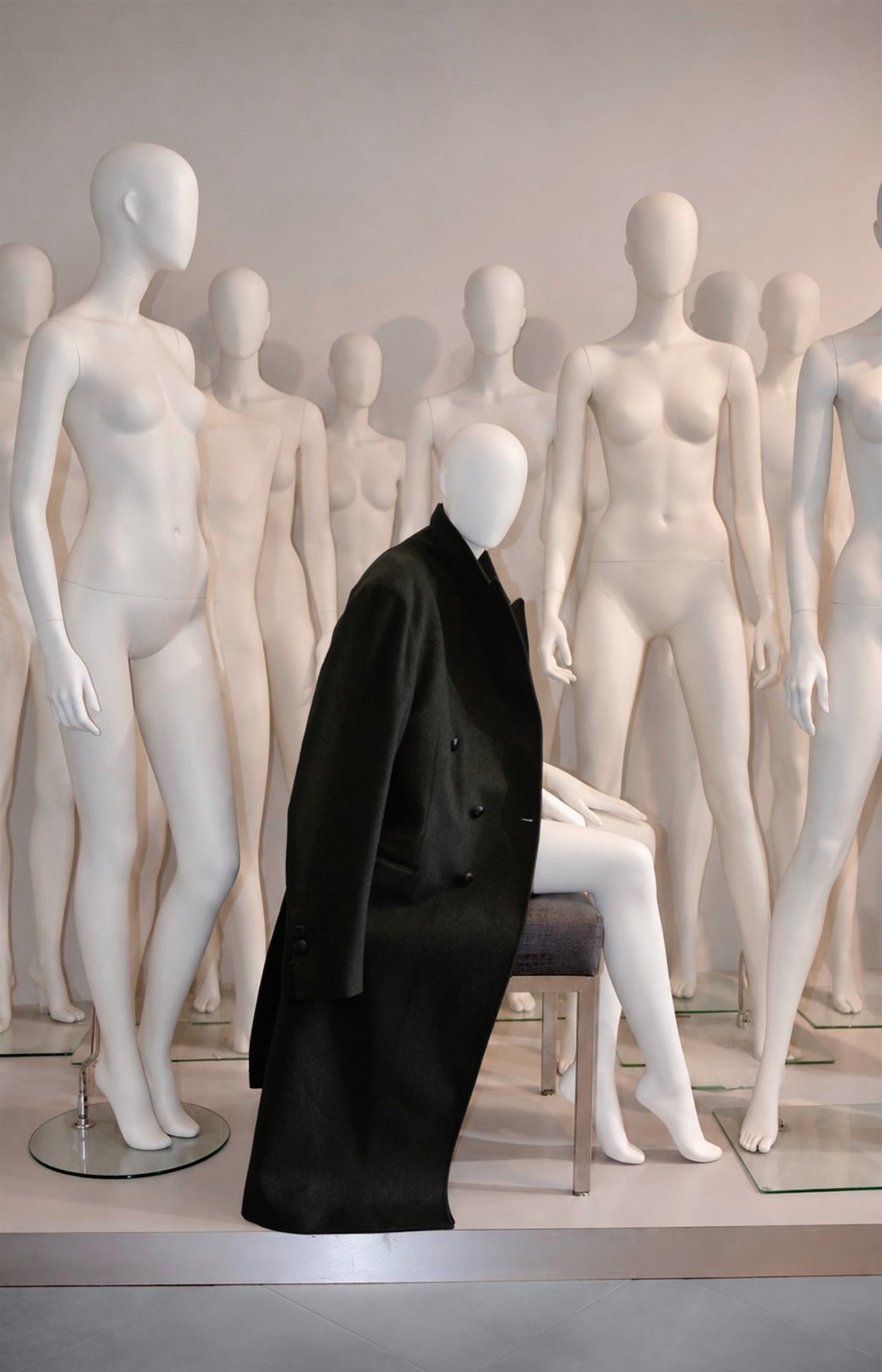 a dark double-breasted loden coat over a seated mannequin, surrounded by white mannequins