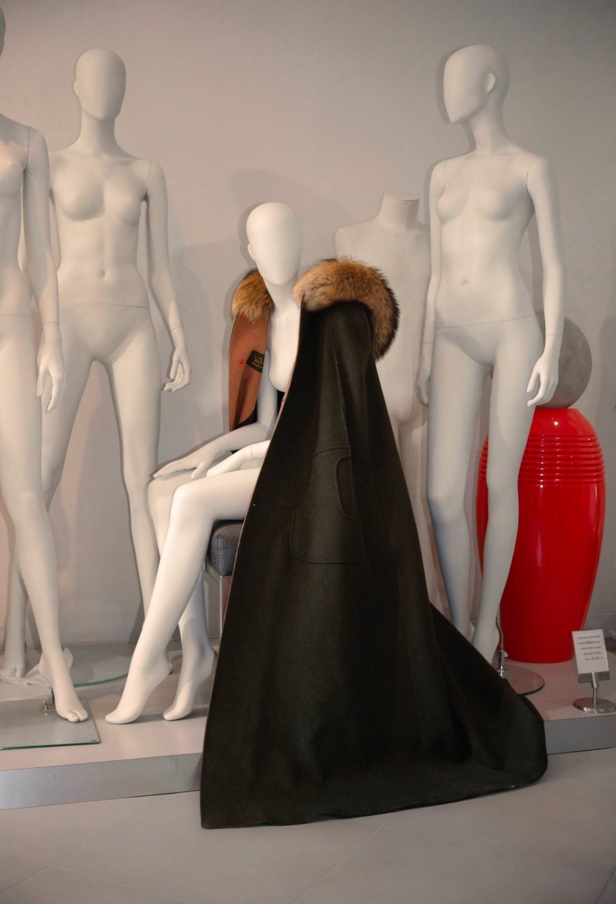 a green loden cape with a fur collar and camel lining on a seated mannequin, white mannequins and a red vase behind