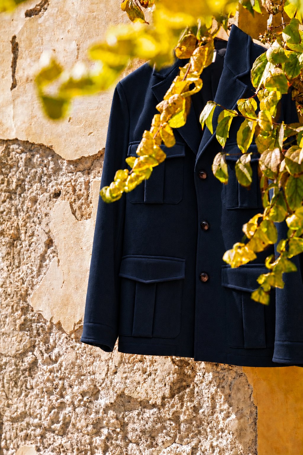 a navy four-pocket jacket hanging against a plaster wall behind yellow leaves