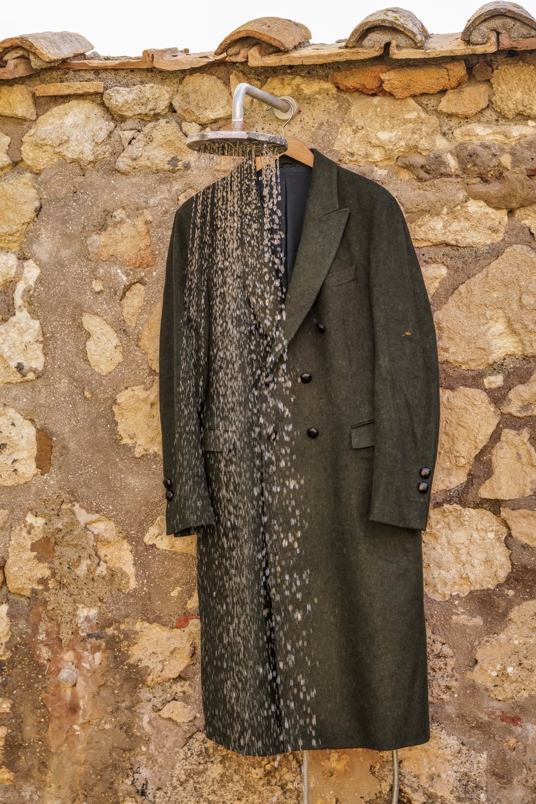 a green double-breasted loden coat under an outdoor shower, the water running off the cloth